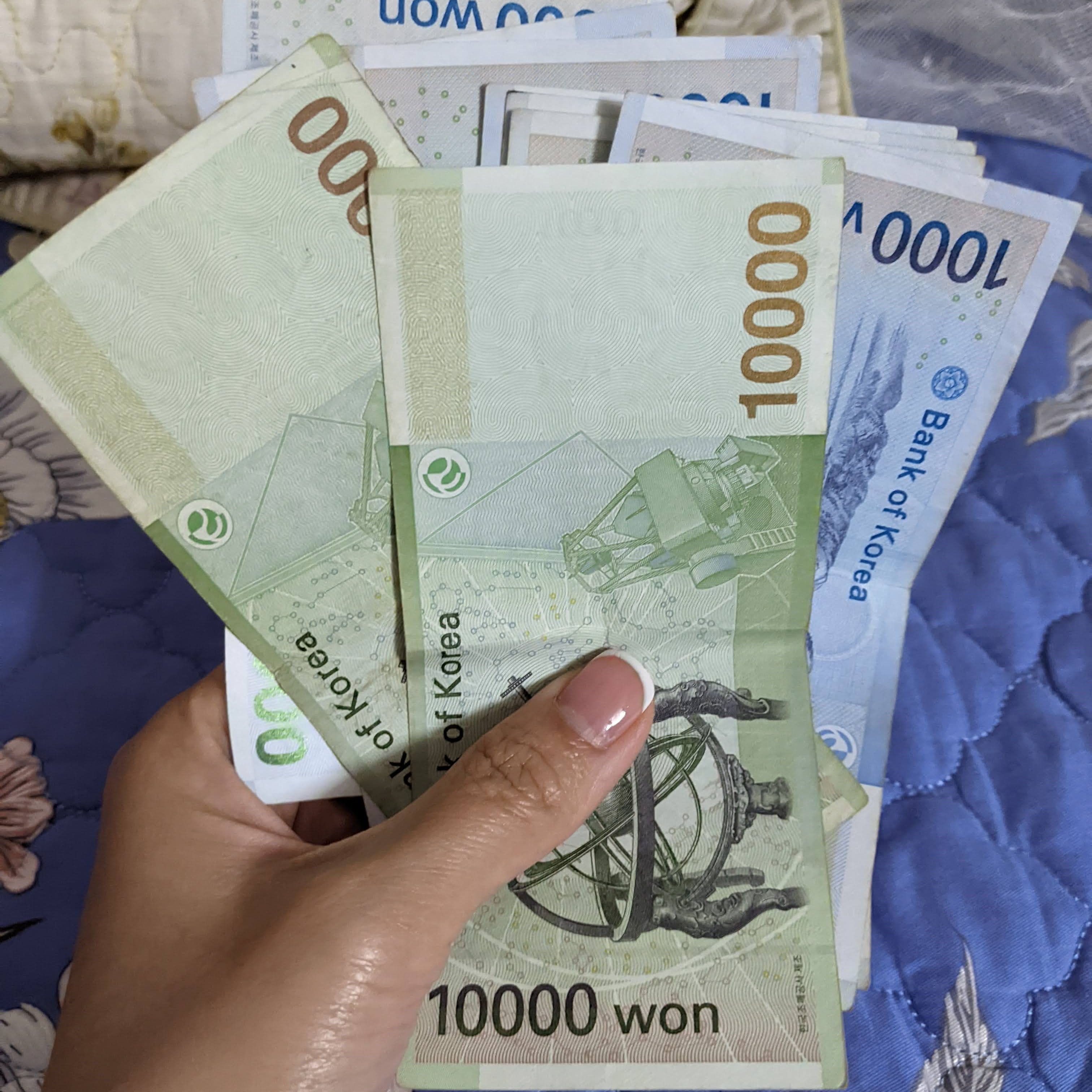 A person holding several South Korean won banknotes, including 10,000 won bills, against a textured background.