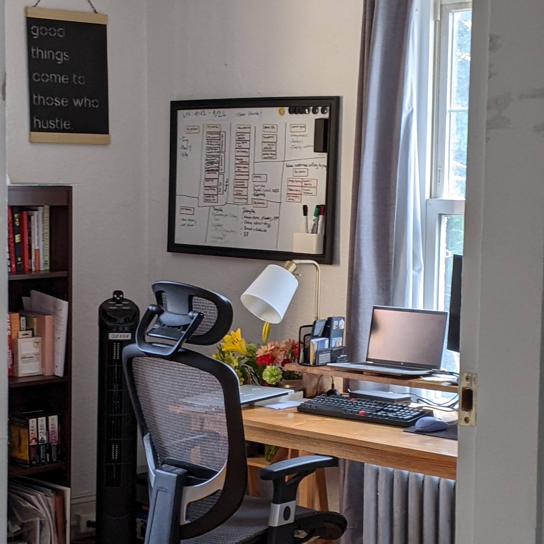 A home office setup with a desk, ergonomic chair, and a laptop. A whiteboard with organizational notes and a lamp are visible. Flowers are placed on the desk, and a bookshelf filled with books is nearby.