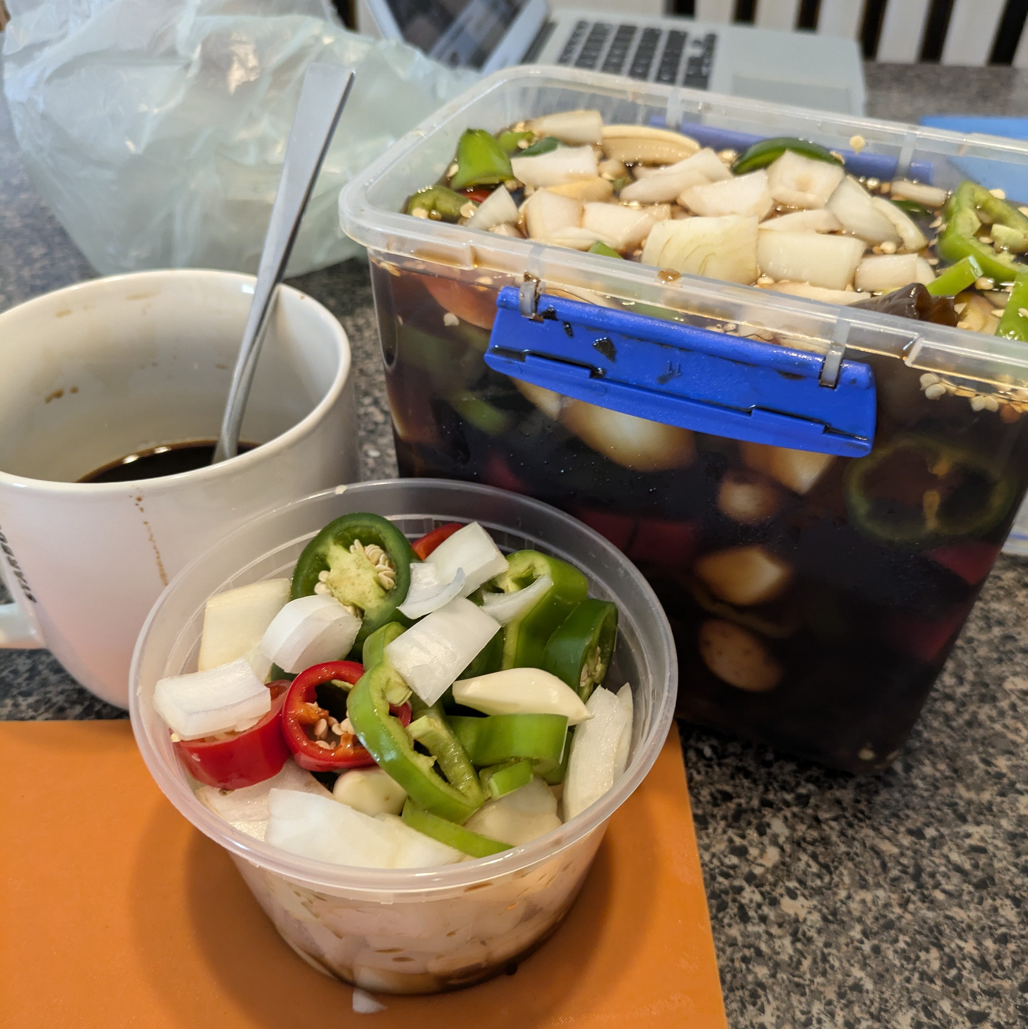 A container filled with marinated vegetables, including sliced onions, green and red peppers, alongside a bowl of fresh chopped vegetables and a cup of dark beverage.
