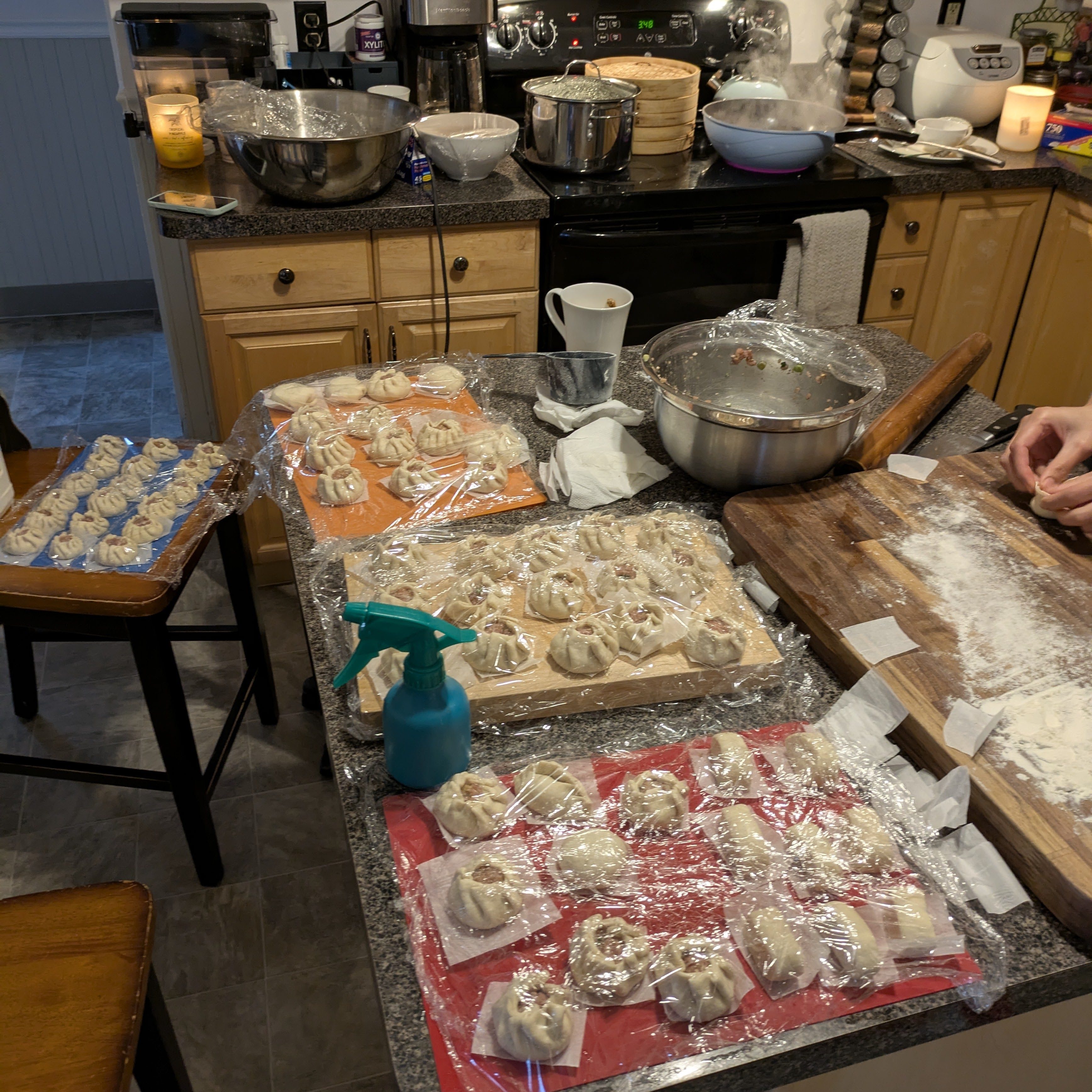 A kitchen countertop filled with various plates of steamed buns, some covered with plastic wrap, and a bowl of dough preparation. There are utensils and containers visible, creating a busy cooking scene.