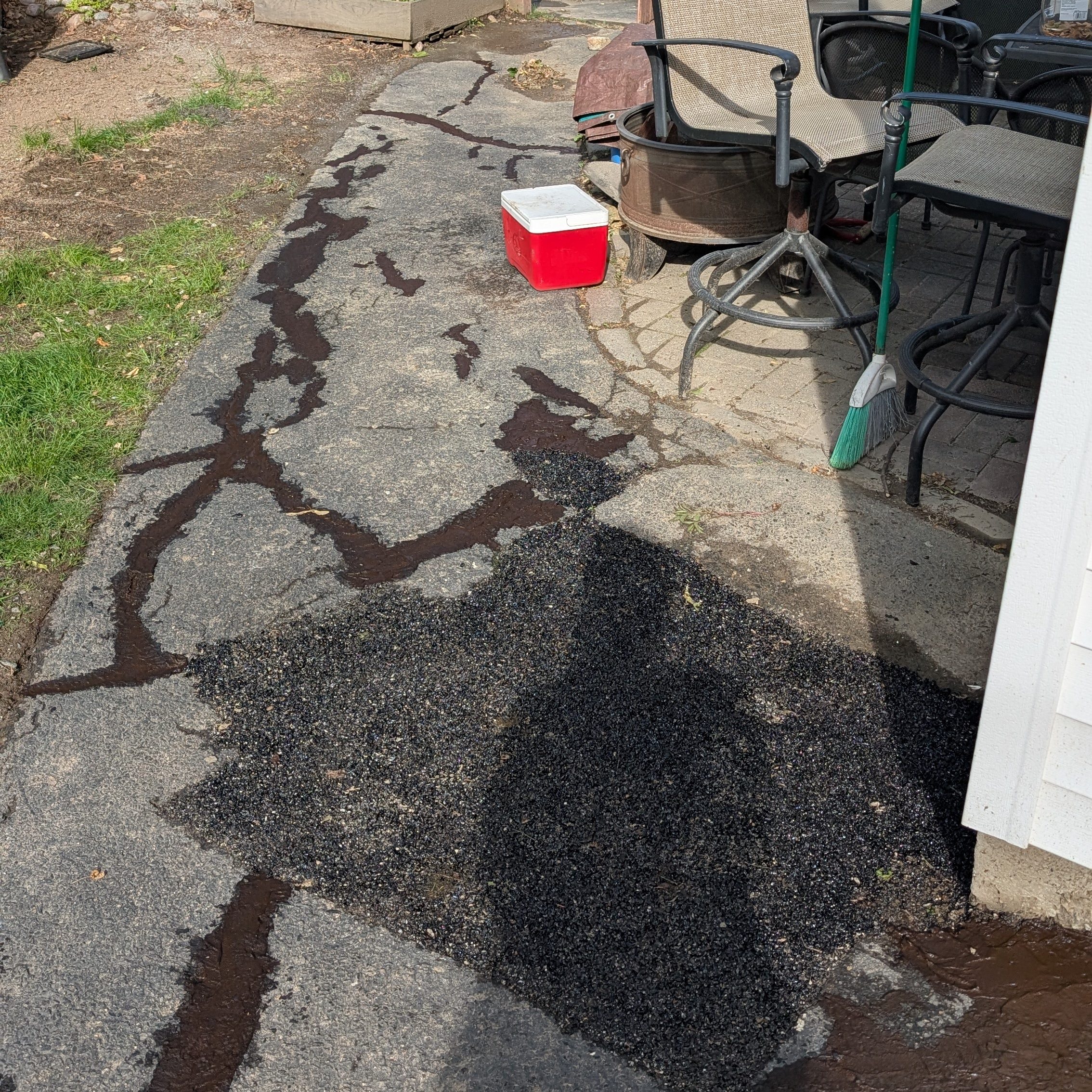 A partially repaired driveway showing cracks and patches of new asphalt next to a patio area with outdoor furniture and a cooler.
