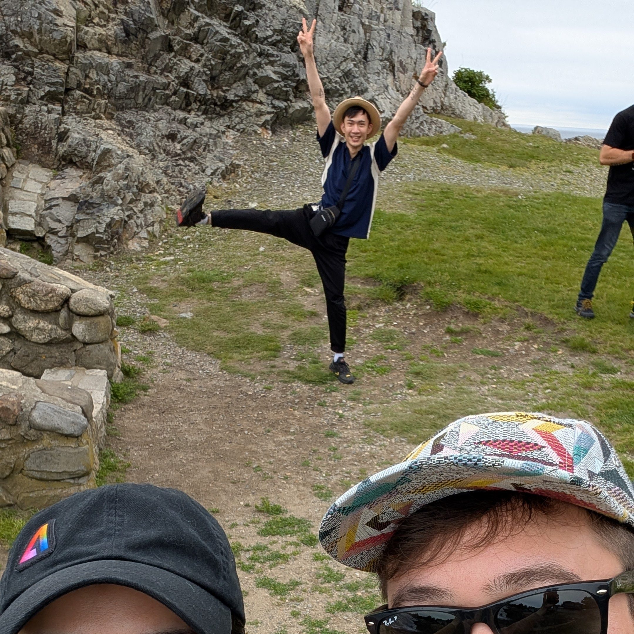 A group selfie at a scenic outdoor location, featuring three individuals, one playfully posing with a peace sign and raised leg.